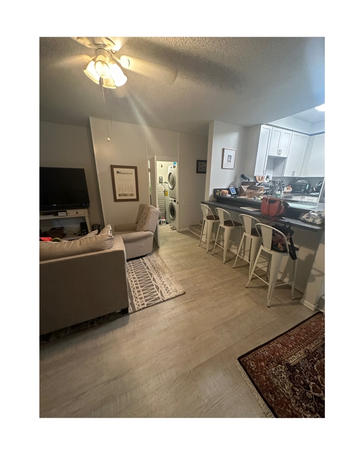 A wide-angle before photo showing an open-concept living area leading into a kitchen. Four white metal barstools sit lined up along a dark raised breakfast counter. In the foreground and left side, a neutral sofa, armchair, pattern rug, light wood-look vinyl flooring, and television setup are visible. In the background, white kitchen cabinets frame the kitchen area next to a small laundry closet with stacked washer and dryer units.